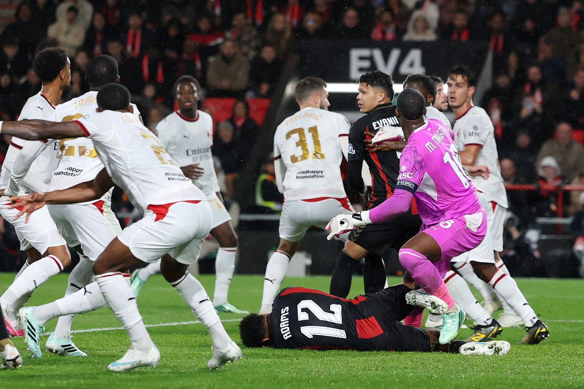 A Bournemouth player wearing number 12 lies on the pitch while a goalkeeper in pink and AC Milan players in white stand around him in the penalty area.