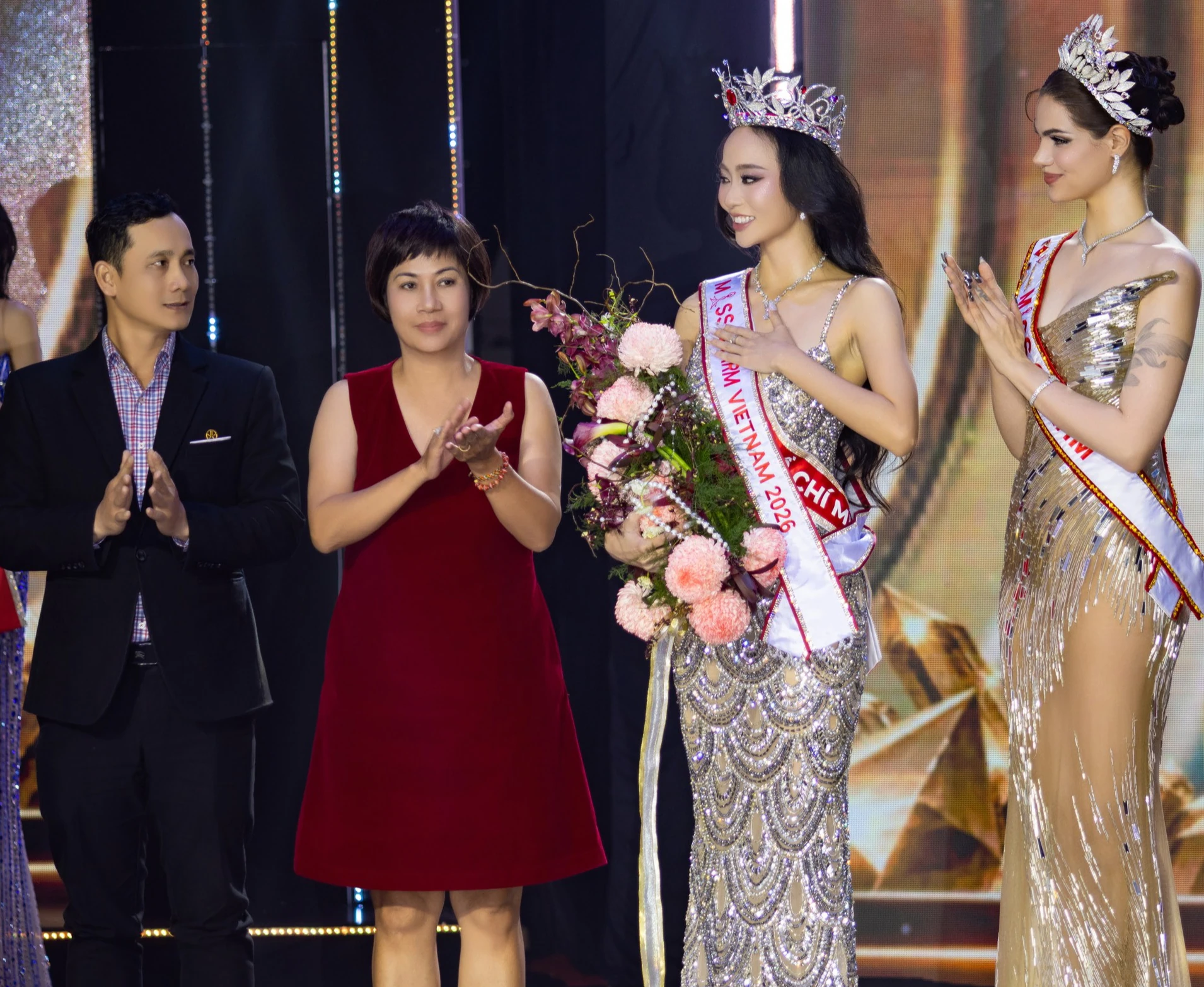 Nguyễn Ngọc Trà My đội vương miện, đeo sash Miss Charm Vietnam 2026, ôm bó hoa trên sân khấu trong khoảnh khắc đăng quang, có người vỗ tay xung quanh.