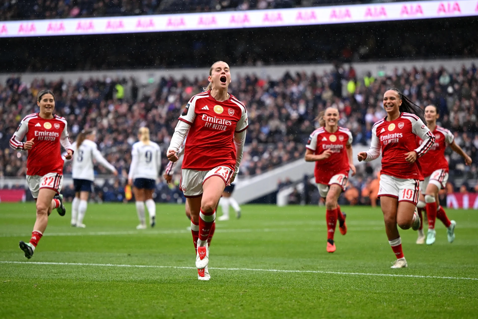 Alessia Russo (số 23) của Arsenal hét lớn ăn mừng, các đồng đội áo đỏ chạy theo trên sân dưới mưa, khán đài kín người