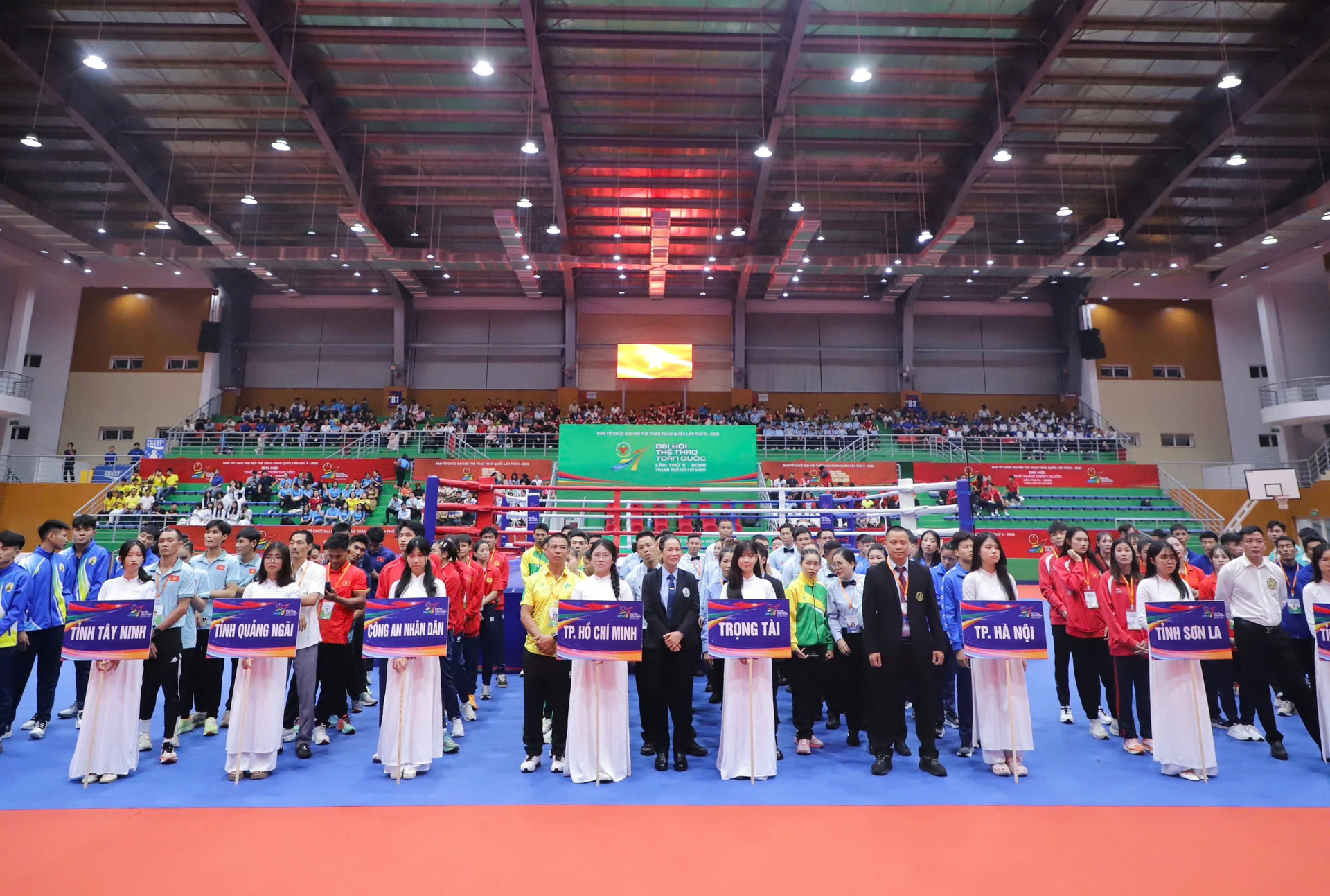 Các đoàn vận động viên, trọng tài và người cầm biển tên đơn vị đứng thành hàng trước võ đài trong nhà thi đấu, trên khán đài có khán giả.
