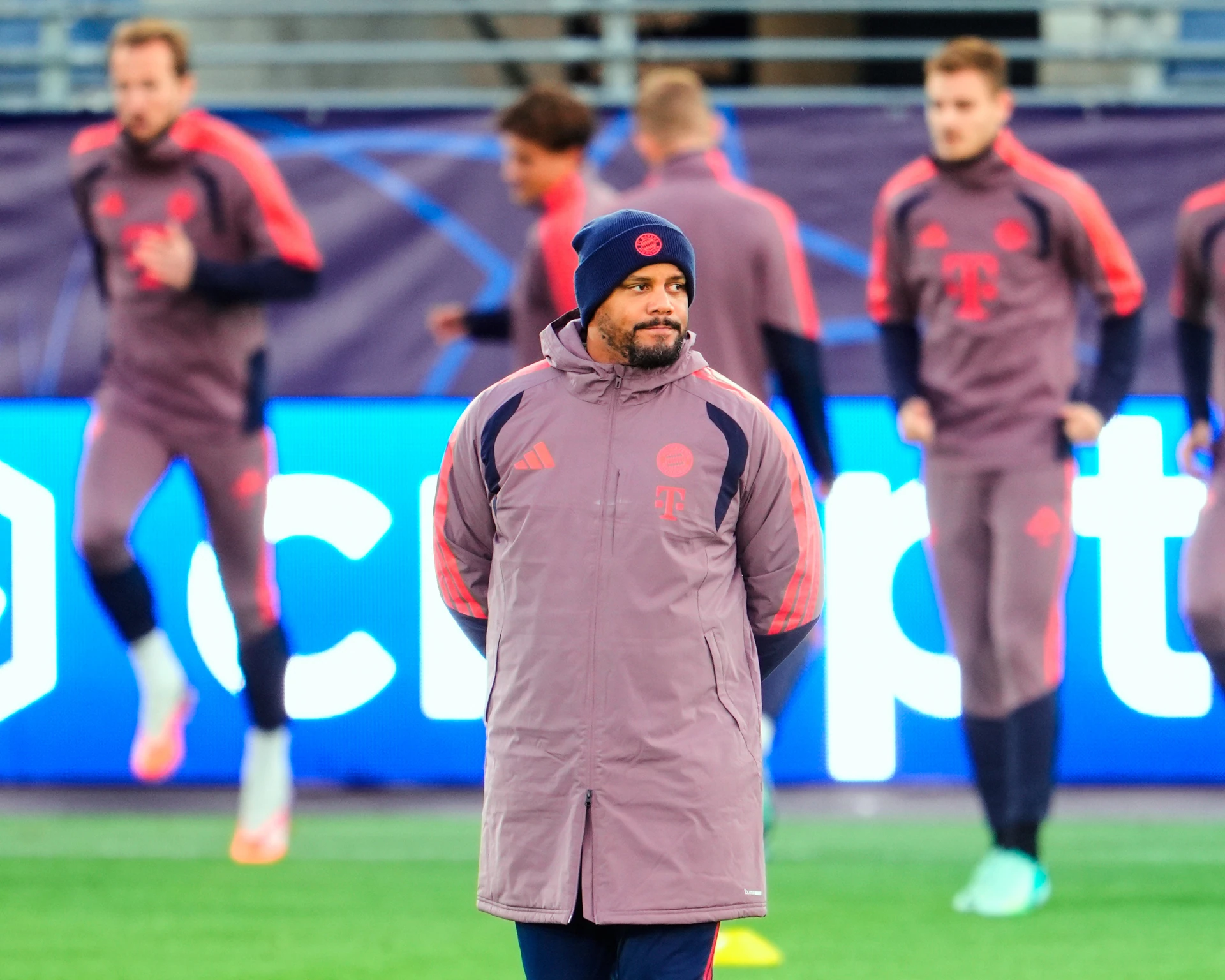 HLV Vincent Kompany đội mũ len, mặc áo khoác dài Bayern Munich đứng chắp tay sau lưng, phía sau là các cầu thủ đang khởi động