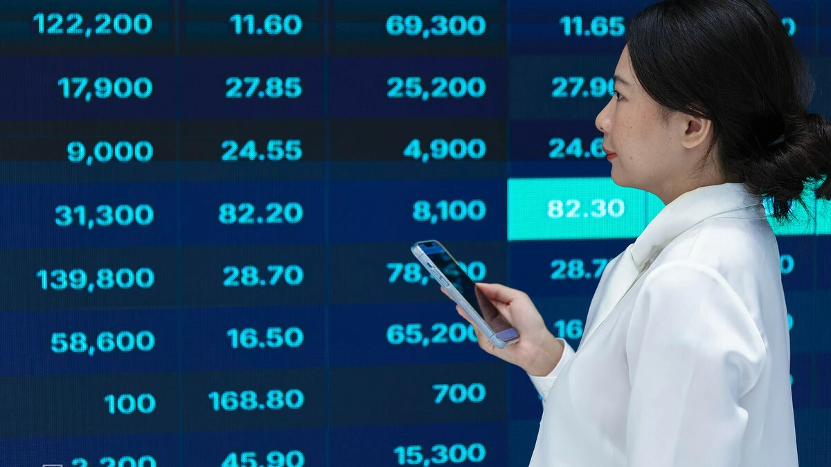 Ảnh minh họa: một phụ nữ cầm điện thoại đứng trước bảng điện giá chứng khoán.