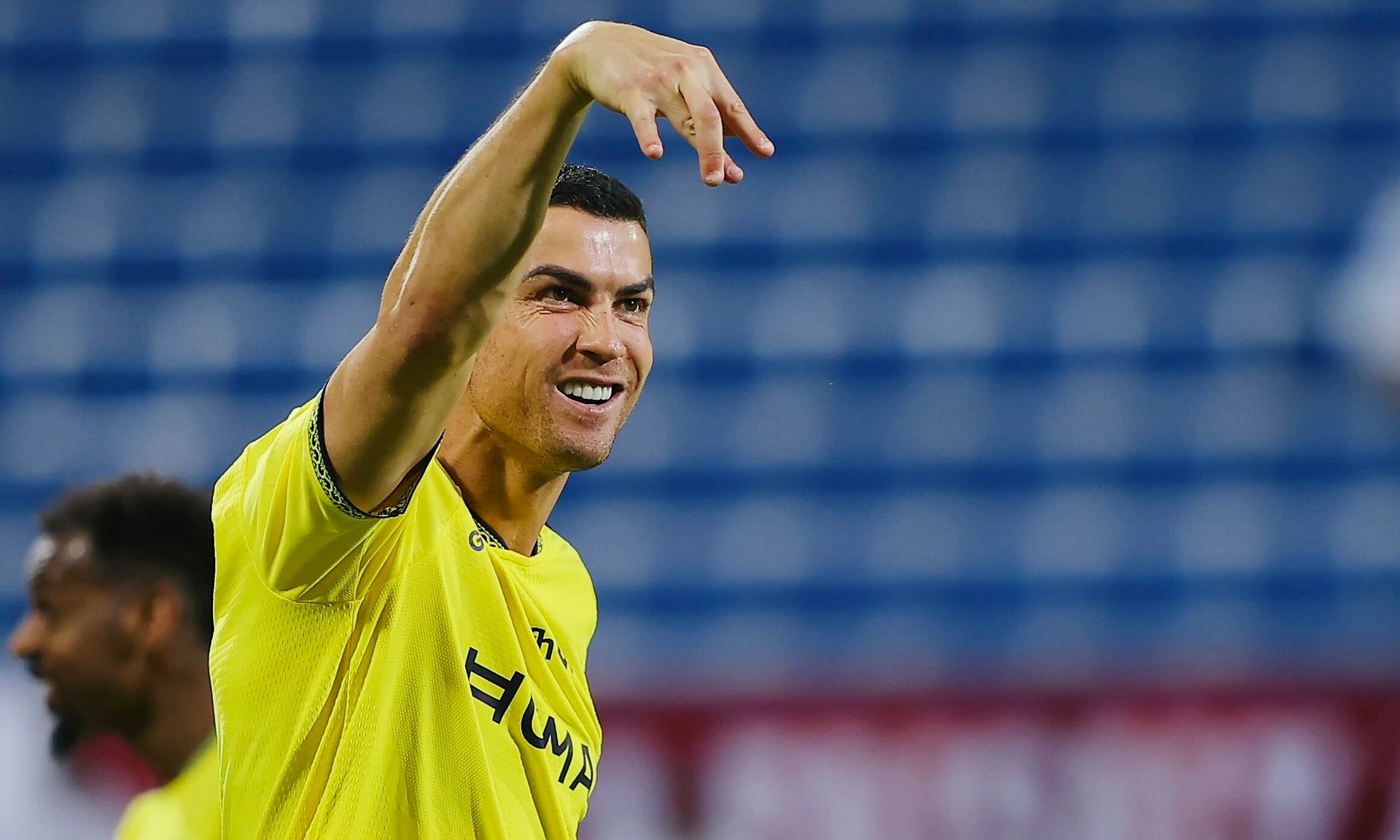 Cristiano Ronaldo trong áo vàng Al Nassr mỉm cười, giơ tay ăn mừng trên sân, phía sau là khán đài ghế xanh