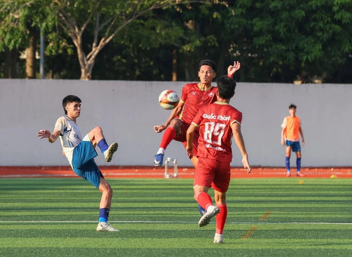 A player in red and a player in white and blue contest a high ball on an artificial pitch, with a white wall and trees behind