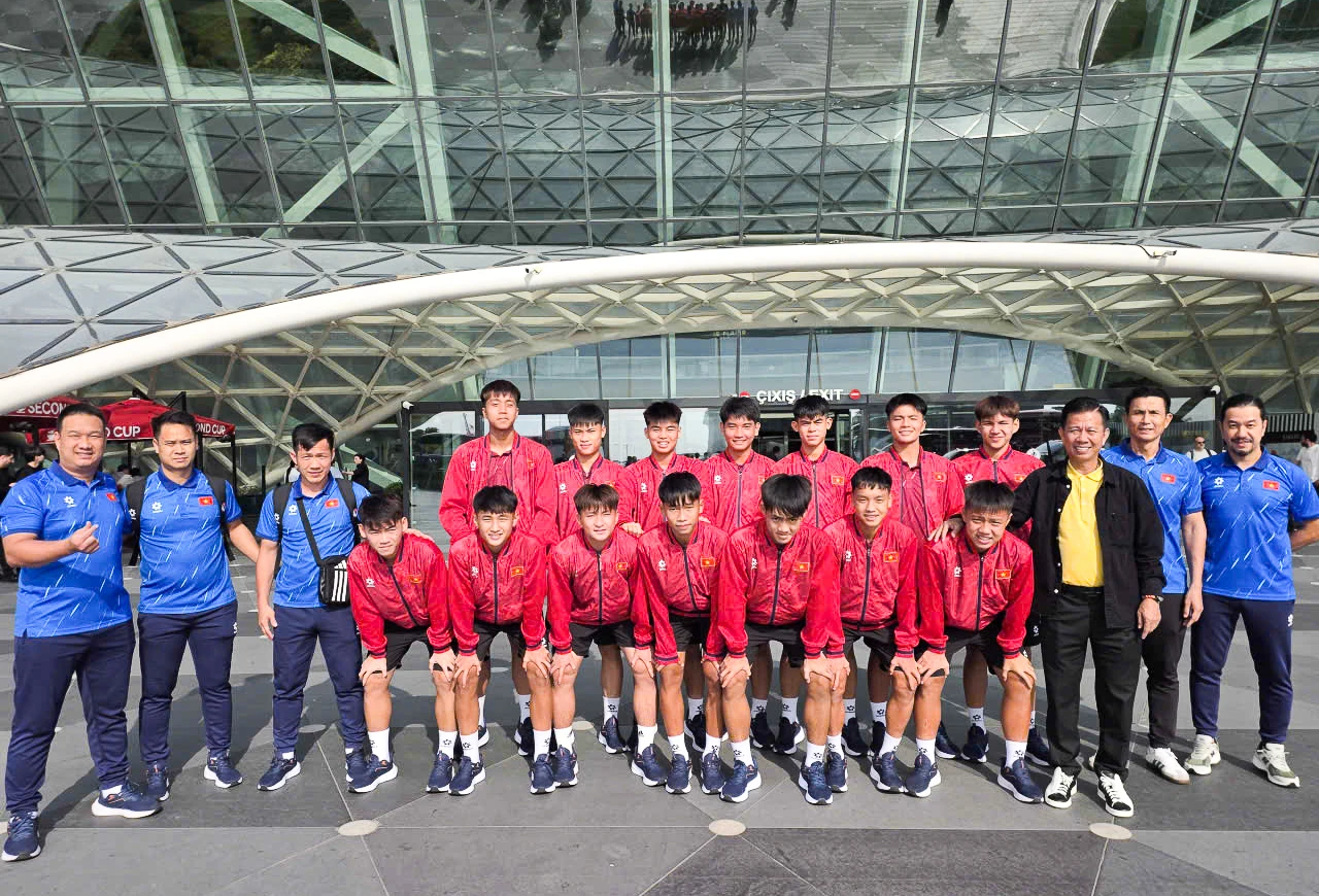 U15 Vietnam players in red jackets pose with their coaching staff in front of a glass-roofed building in Baku, Azerbaijan.