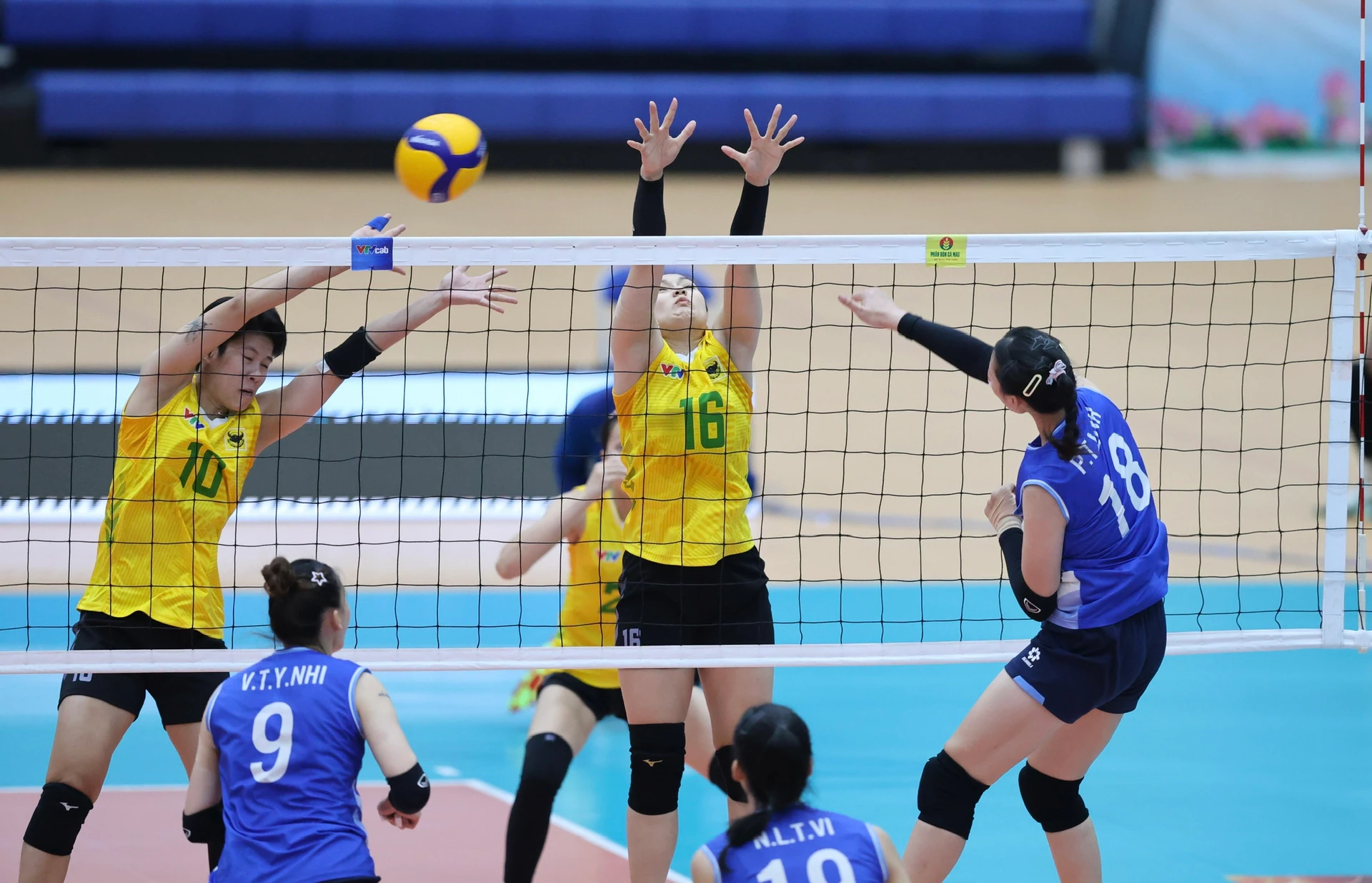 Two women players in yellow jump to block at the net as a player in blue spikes the ball during a women's volleyball match.