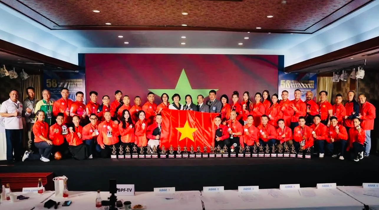 Các thành viên đội tuyển thể hình Việt Nam mặc áo đỏ chụp ảnh tập thể cùng cờ đỏ sao vàng và nhiều cúp trên sân khấu