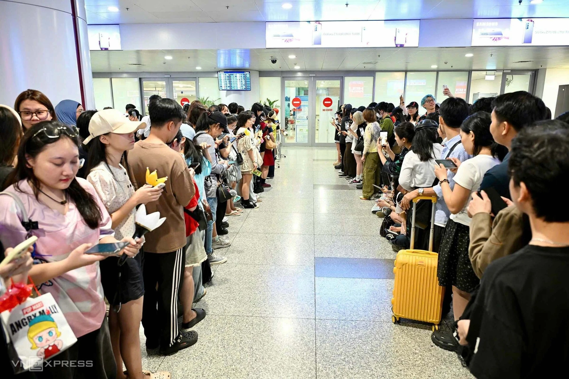 Long lines of fans stand on both sides of a walkway in an airport hall, many holding phones and light sticks, with a yellow suitcase on the right.