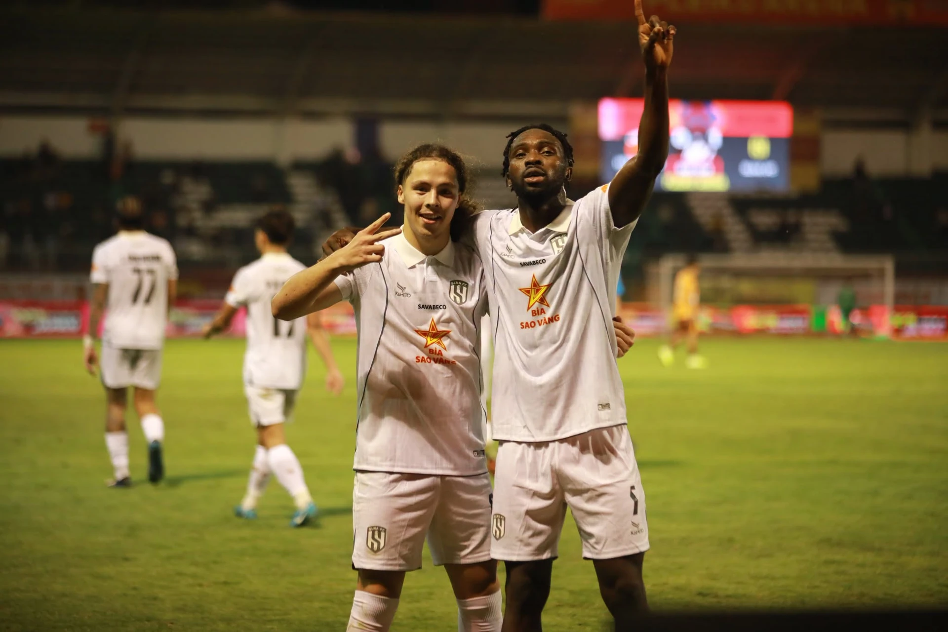 Two SLNA players in white kits celebrate with arms around each other on the pitch, one holding up two fingers and the other pointing to the sky.