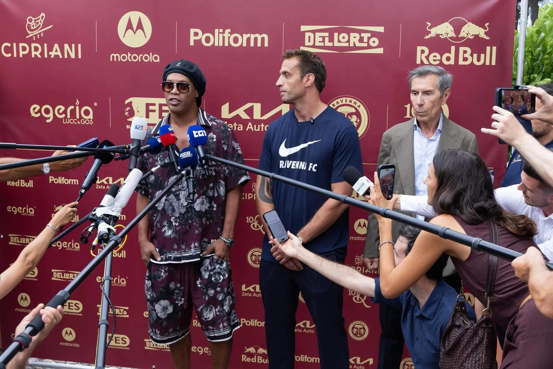 Ronaldinho đội mũ nồi, đeo kính râm trả lời phóng viên trước tấm phông Ravenna, bên cạnh là hai người đàn ông
