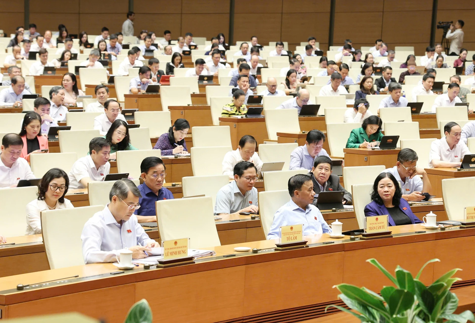 Các đại biểu Quốc hội ngồi theo hàng trong hội trường, nhiều người ghi chép hoặc dùng máy tính bảng, trước mỗi người có biển tên.